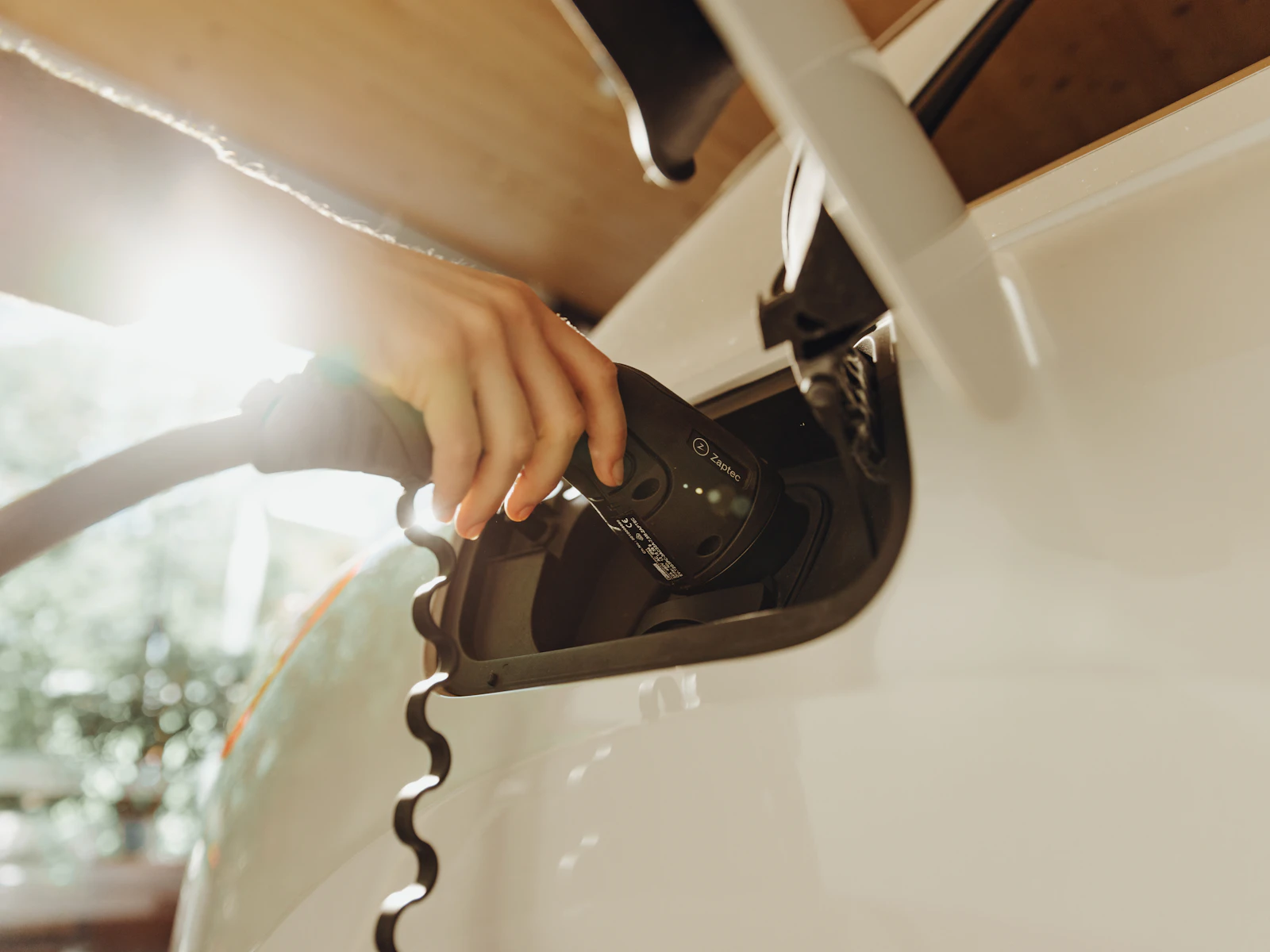 A hand connecting a charging cable to an electric vehicle in sunlight